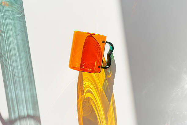 istock Orange Glass Mug with bright sunlight Shadows, transparent mug casting dynamic, vibrant lights reflections and shadows on light white surface, creative aesthetic top view, minimal close up 2210967868