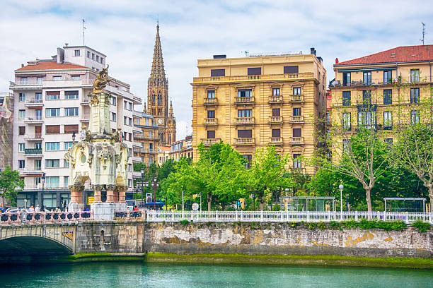 Sunlight casts soft reflections on a quiet river in Bilbao’s old quarter, where a historic church and stone bridge meet colorful urban life and calm skies