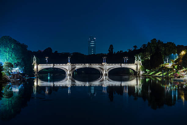 Night view of the Umberto I Bridge in Turin, Italy, lit up and reflecting on the Po River, with calm waters and a clear sky.