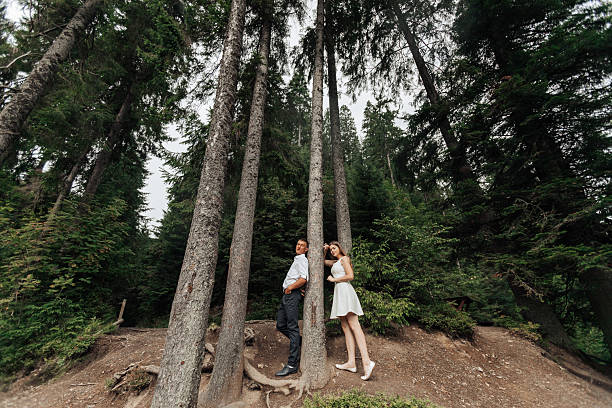 A couple is posing in front of a tree in a forest. The man is wearing a white shirt and the woman is wearing a white dress. Scene is romantic and peaceful