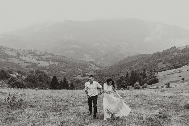 A couple is running through a field with mountains in the background. The man is wearing a white shirt and jeans, while the woman is wearing a dress. The scene is peaceful and romantic