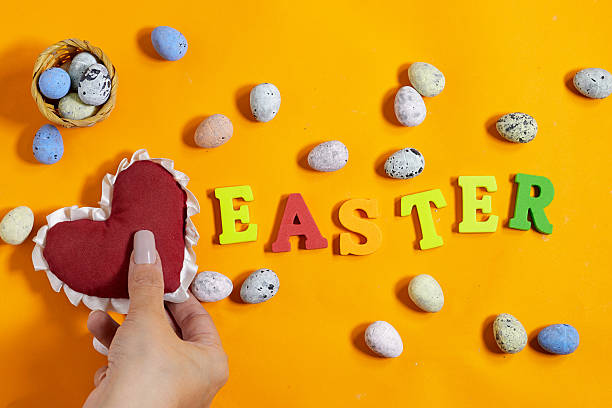 istock Vibrant Easter-themed arrangement featuring a hand holding heart-shaped decoration colored eggs and the word Easter on an orange backdrop evoking festivity and warmth 2211015300
