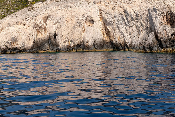 A rugged limestone cliff meets the calm blue sea along the coastline of a Greek island under soft sunlight.