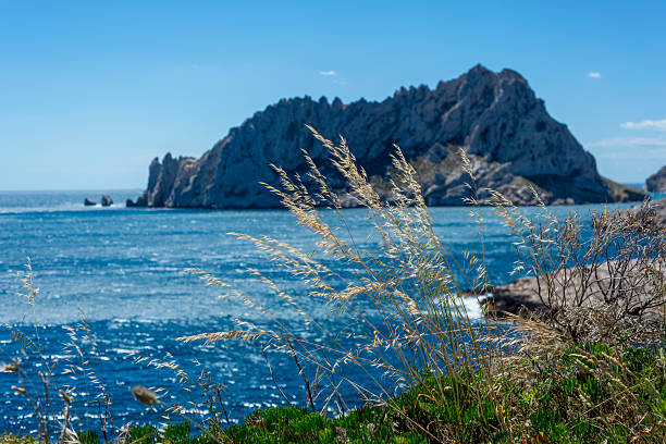 Bright sunny day with clear blue sea and rocky coastline. Sailboat anchored near shore with green hills in the background. Scenic coastal landscape with calm ocean water and natural rocks.