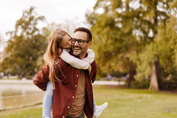 Happy Loving Family! Cute Little Daughter Kissing Her Happy Father While Giving Piggyback Ride in the City Park. Having Fun Together.