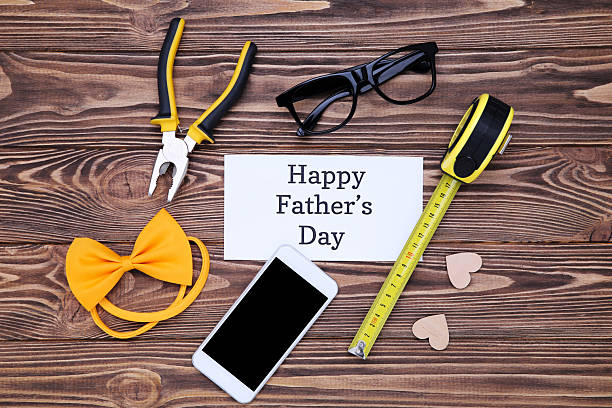 istock Text Happy Father's Day with instruments, smartphone, eyeglasses and bow tie on wooden table 2211023260