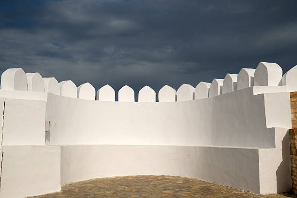 A tall white wall with pointed battlements stands solidly, contrasting with dark clouds overhead. The scene evokes a sense of history and strength in this architectural marvel.