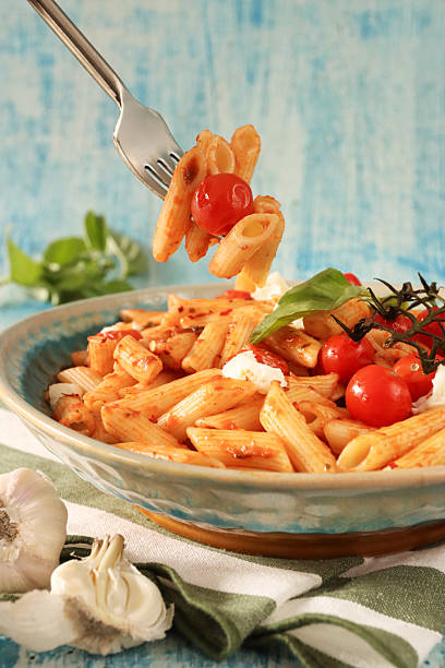 Stock photo features a fork lifting penna pasta from a beautifully arranged dish, topped with juicy cherry tomatoes, fresh basil leaves, creamy burrata cheese, and tender buffalo mozzarella balls. The vibrant colors and textures highlight the freshness of this Italian-inspired meal