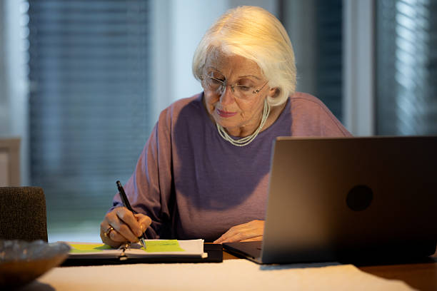 istock Senior Caucasian Woman Writing Notes While Using Laptop in Cozy Indoor Setting 2211038077