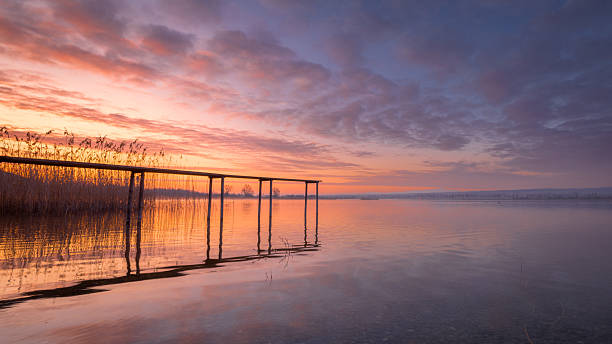 A serene sunrise over Lake Constance with a wooden pier extending into the calm water. The sky transitions from soft pink to deep blue, reflecting on the lake’s surface.