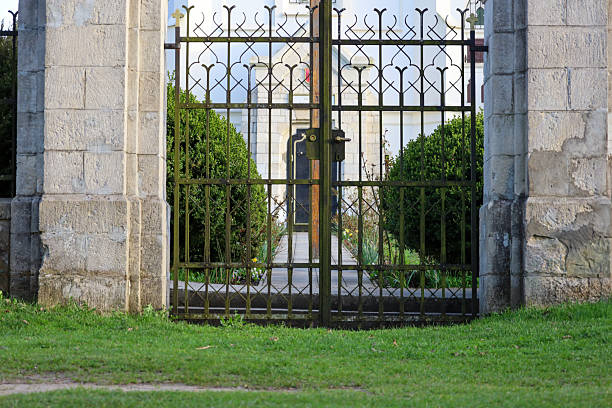 Calgary, Alberta, Canada- July 31, 2025:  High black wrought iron security and privacy gate in a non urban area.  One side open. Large green area with some evergreen trees.  Blue sky.
