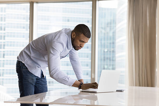istock Young African businessman use laptop standing leaned over workplace desk 2211065036