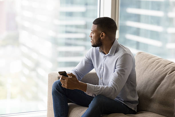 istock African man resting on couch holds smartphone staring out window 2211065744