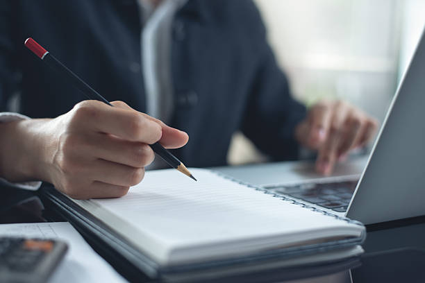 istock Clsoe up, business woman hand with pencil writing on paper notebook, working on laptop computer at modern office. Businesswoman taking notes, planning business project, doing finance, business process 2211071632