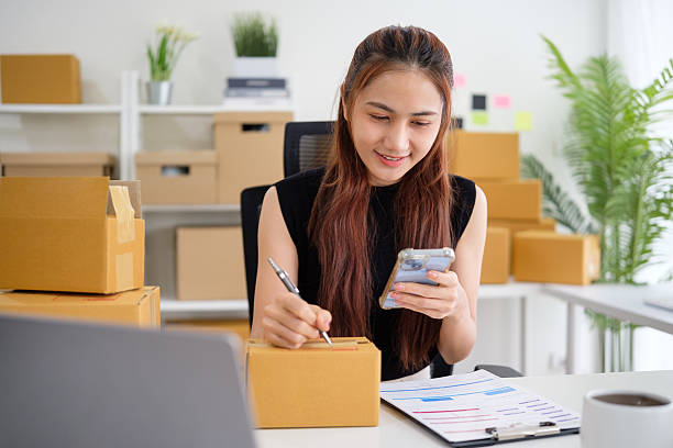 istock Small business entrepreneur using her phone while preparing packages for delivery. Small business operations and e-commerc concept. 2211077453
