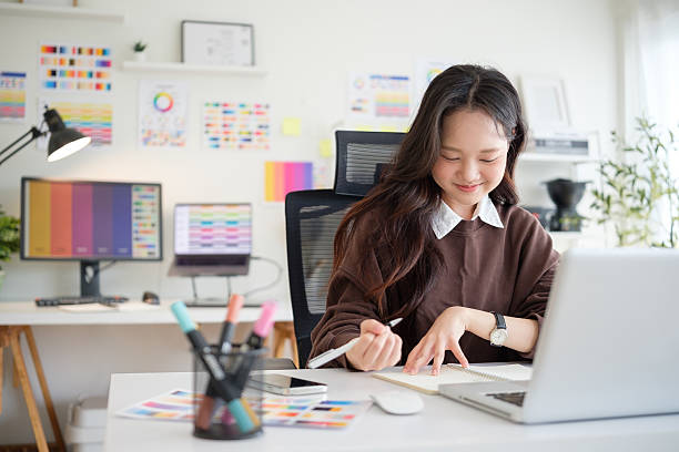 istock Young female graphic designer jotting down ideas in a notebook while working on a laptop at creative workdesk 2211077674