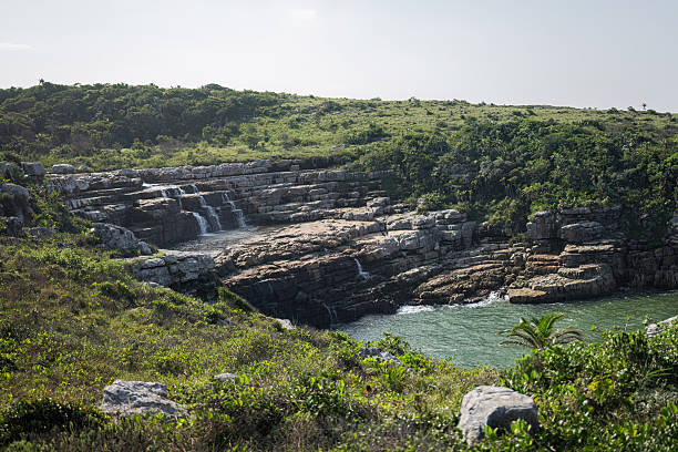 A dramatic, multi-level waterfall cascades down layered rock into a secluded cove below, surrounded by lush vegetation and rugged cliffs. This photo was taken in the Wild Coast of the Eastern Cape, South Africa.