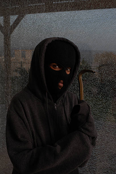 A masked person wearing a dark hoodie stares intently at the camera while holding a crowbar, standing before a large, shattered glass window. The moody lighting and composition convey a sense of mystery, tension, and criminal intent. This photo is ideal for illustrating themes related to burglary, crime, urban danger, and law enforcement scenarios.