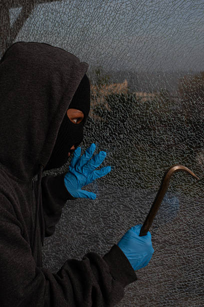 A masked individual in a dark hoodie and blue gloves examines a shattered glass window while holding a crowbar. The dramatic lighting, visible tension, and close-up detail suggest a moment of decision in an attempted break-in or criminal act. This image is ideal for illustrating themes of burglary, law enforcement, and home security breaches.