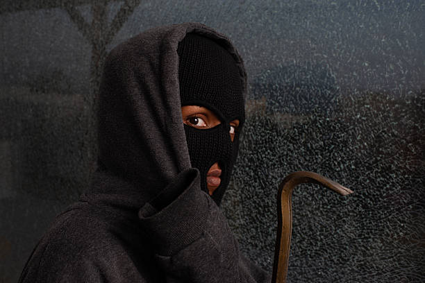 A person in a dark hoodie and black ski mask looks intensely at the camera while partially concealing their face and holding a crowbar. The shattered glass background and dramatic lighting emphasize themes of intrusion, secrecy, and criminal activity. This photo is suitable for crime, law enforcement, and urban security stories.