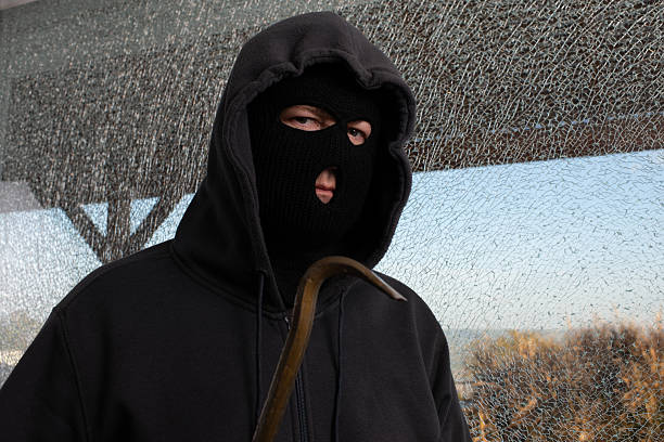 A person dressed in a black hoodie and ski mask holds a crowbar while standing in front of a shattered window. The cold expression and urban setting convey tension, threat, and unlawful intent. This image is suited for illustrating crime, burglary, and urban safety concerns.