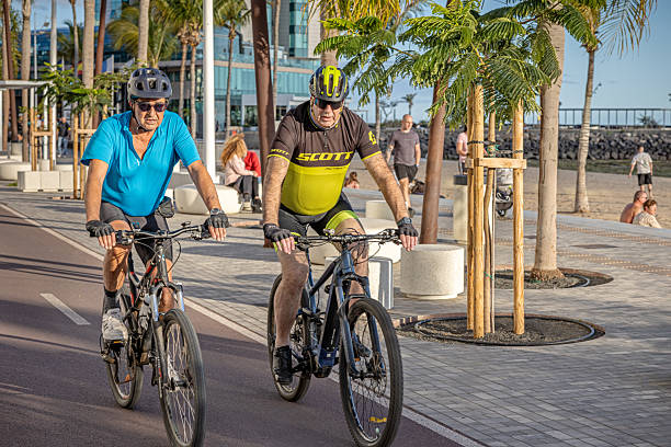Arrecife, Lanzarote, Canary Islands, Spain - March 11th 2025:  This image captures a middle-aged couple enjoying a scenic bike ride along the beach promenade in Arrecife, Lanzarote. Dressed in sporty cycling attire, they pedal leisurely past palm-lined walkways, with the ocean stretching into the horizon. The relaxed yet energetic atmosphere reflects the island’s appeal for outdoor activities, where locals and tourists alike embrace the coastal breeze and stunning views.