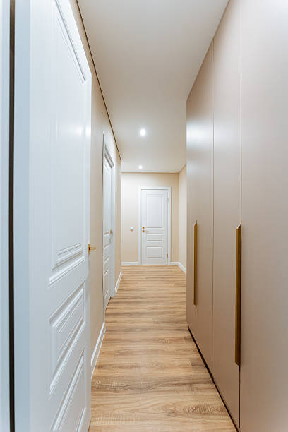 In this expansive and visually appealing long hallway, one can observe the beautiful wooden floors that add warmth and character, complemented by a series of pristine white doors
