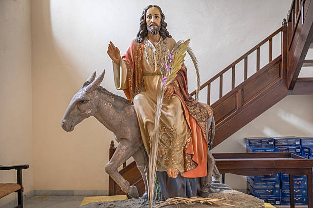 Parroquia Matriz de Nuestra Señora de Guadalupe, Teguise, Lanzarote, Canary Islands, Spain - March 4th 2025:  This detailed image captures a figurine of Christ riding a donkey, adorned in an ornate robe and cape, holding a bouquet of palm branches. The craftsmanship highlights the movement of the donkey, reinforcing the dynamic nature of the scene. Positioned indoors, the figure stands in front of a staircase, with stacked boxes in the background, suggesting a setting related to religious preparation or storage. The representation likely symbolizes Palm Sunday, commemorating Christ’s entry into Jerusalem.