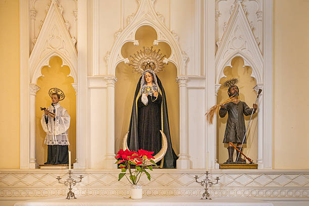 Parroquia Matriz de Nuestra Señora de Guadalupe, Teguise, Lanzarote, Canary Islands, Spain - March 4th 2025:  This image captures a beautifully adorned Catholic altar featuring three distinct saint figures, each representing a revered aspect of faith and devotion. The central figure, dressed in black robes with a golden crown and halo, holds a rosary, symbolizing deep spiritual contemplation. To the left, a saint in white and black clerical garments holds a cross, signifying religious guidance and protection. On the right, another saint, dressed in dark robes, carries a bundle of wheat and a staff, possibly representing sacrifice and sustenance. The altar is elegantly framed by intricate architectural details, with a vase of red flowers placed in front of the central figure, enhancing the solemn and sacred atmosphere.