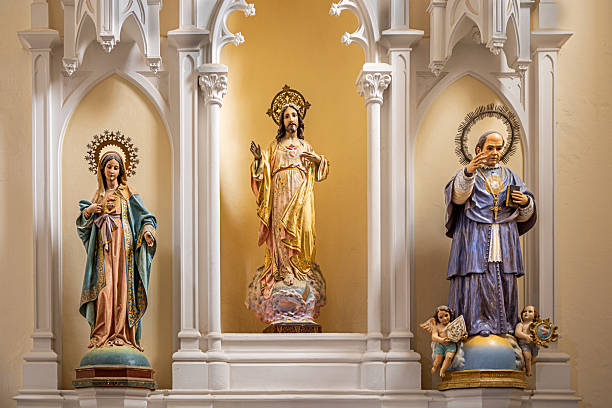 Parroquia Matriz de Nuestra Señora de Guadalupe, Teguise, Lanzarote, Canary Islands, Spain - March 4th 2025:  This image showcases a beautifully crafted Catholic altar featuring three distinct saint figures, each placed within individual niches. The leftmost figure is dressed in blue and pink robes, adorned with a halo decorated with star-shaped embellishments. The central figure, draped in golden garments, stands upon a cloud, with a floral-adorned halo emphasizing its divine presence. The rightmost saint, dressed in blue and white robes, holds a book and wears a radiant halo with sunburst-like details. At the feet of the rightmost figure, two small angels add to the spiritual depth of the scene. The intricate carvings and rich symbolism reflect the deep religious tradition and artistic craftsmanship central to Catholic worship.