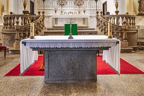 Parroquia Matriz de Nuestra Señora de Guadalupe, Teguise, Lanzarote, Canary Islands, Spain - March 4th 2025:  This image captures a solemn religious setting featuring a table positioned in front of a richly adorned Catholic altar. The table, draped in a pristine white cloth, holds two elegant candlesticks and a crucifix, reinforcing its ceremonial significance. Behind it, the altar showcases intricate religious statues and sacred figures, framed by detailed architectural elements that enhance the spiritual ambiance. The deep red carpeting adds warmth to the scene, while the high, vaulted ceiling emphasizes the grandeur of the church interior.