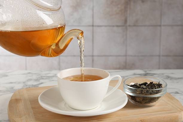 Pouring refreshing green tea from teapot into cup at white marble table, closeup
