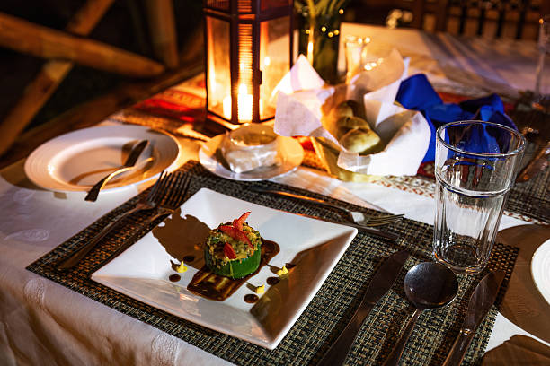 A professionally plated gourmet appetizer is set on an outdoor table with cutlery, glassware, and a candle-lit lantern. The scene is prepared for fine dining, captured in warm artificial light against a wooden lodge backdrop. This photo was taken in Uganda.
