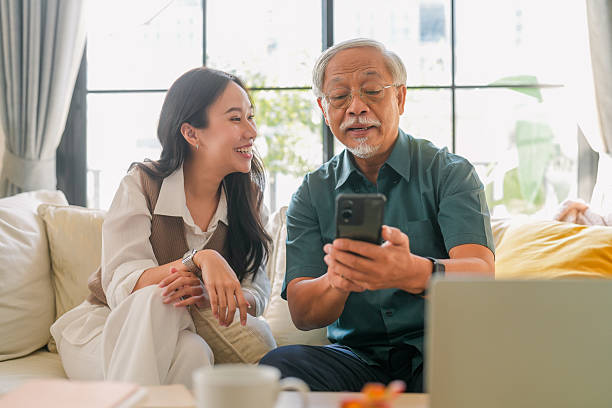 Asian senior father takes help in his smartphone, regarding a new application based on the latest technology Young woman daughter teach help in smartphone apllication use