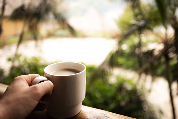 A hand holds a white ceramic mug over a wooden balcony railing with a blurred view of green vegetation, palm trees, and thatched rooftops in the background. This photo was taken in Uganda.