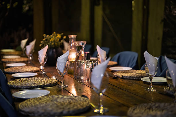A long wooden dining table is neatly arranged with plates, glasses, napkins, and cutlery. Candles in the center provide warm evening lighting for a prepared dinner setting.