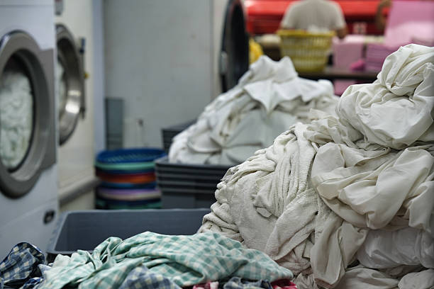 Piles of sorted laundry including white linens and colorful textiles in plastic bins inside a commercial washing plant, with machines and workers in the background.