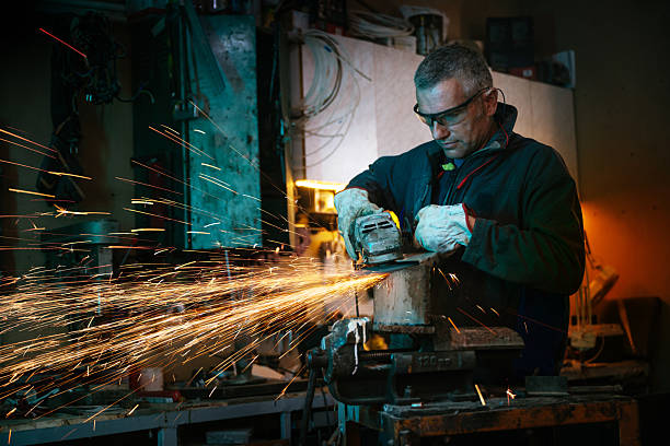 istock Industrial worker in a dimly lit workshop wearing protective goggles and operating an angle grinder with flying sparks around 2211159956