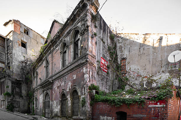 Istanbul, Turkey – November 22, 2015: A street corner surrounded by abandoned and worn-down buildings. The facade of the two-story building in the foreground is crumbling, with walls covered in graffiti and ivy. A red-and-white striped warning sign hangs on the entrance door. In the background, tall and neglected concrete walls stretch along the street.