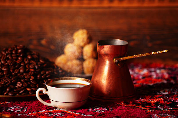 A warm and inviting still life captures the essence of traditional turkish coffee preparation. A classic white porcelain cup with gold trim is filled with dark, rich turkish coffee, with delicate steam rising to indicate its freshness. Beside it sits a gleaming copper cezve, the traditional pot used for brewing. A small mound of roasted coffee beans adds to the aromatic suggestion, all arranged on an intricately patterned red rug. The blurred background hints at a rustic wooden setting, enhancing the authentic and cultural feel of the scene.