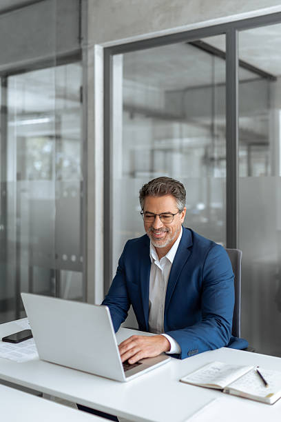 istock Mature Latin business man ceo manager using laptop computer in modern office. Smiling senior businessman entrepreneur sitting at desk working on financial project, looking thinking at screen. Vertical 2211175028
