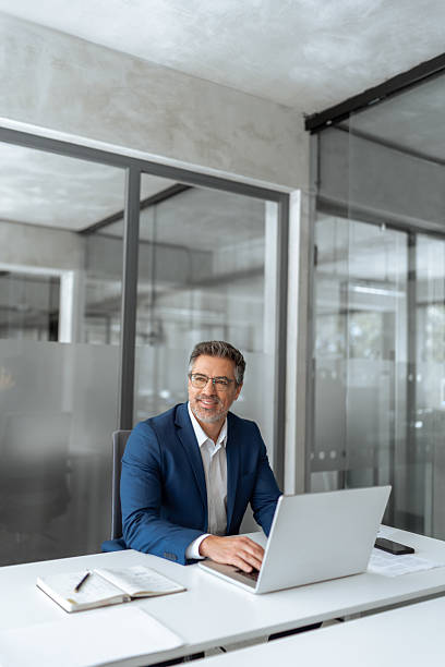 istock Mature Latin business man ceo manager using laptop computer in modern office. Smiling senior businessman entrepreneur sitting at desk working on pc financial project, looking thinking aside. Vertical 2211175031