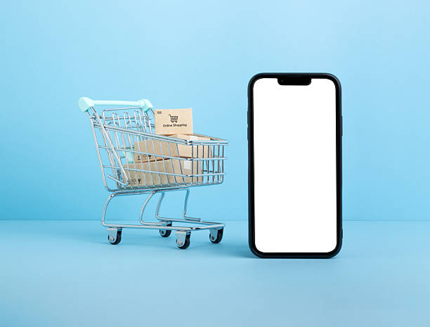 istock Shopping cart filled with small packages placed beside a smartphone with a blank white screen on a blue background, representing online shopping, e-commerce, and digital retail innovation 2211176859