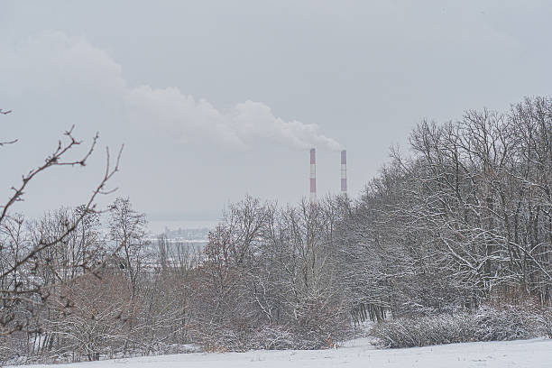 istock A wintry landscape with snow-covered ground, bare trees dusted with snow, an industrial facility or power plant in the distance emitting smoke, and an overcast sky with reduced visibility due to fog 2211183058