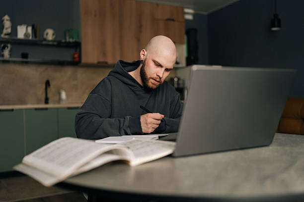 istock Bearded bald young man in hoodie taking notes sitting at kitchen table with laptop computer, managing online business and enjoying comfort of working from home. 2211185509
