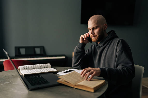 Focused music composer talking on mobile phone and reading book at home studio, with laptop and sheet music on table, working remotely on new project.