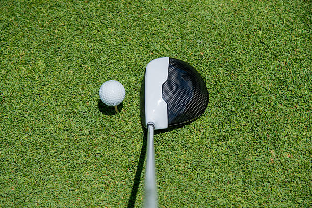 istock golf club driver is positioned behind a golf ball on a tee, ready for a swing, resting on a vibrant green grass surface under bright sunlight. 2211200806