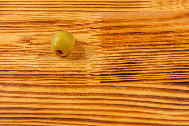 A single green olive is placed on a smooth wooden surface, showcasing the natural grain and texture. The simplicity highlights the olive's color and shape against the wood.