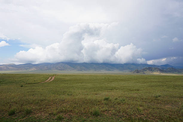 Vast steppe landscape in mongolia showcasing a winding dirt road, distant mountains rising majestically, and a dramatic cloudy sky enhancing the serene atmosphere of the wilderness