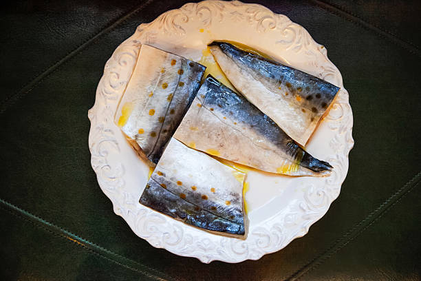 Four fresh mackerel fillets seasoned and drizzled with olive oil, resting on an elegant white plate, prepared for cooking and placed on a dark green leather surface. Concept of pescetarianism and omega 3